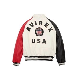 Op de afbeelding: Een varsity-jack met een wit achterpaneel, een rode rechtermouw en een zwarte linkermouw. De achterkant toont de zwarte tekst AVIREX, een ronde patch met een Amerikaans vlag-embleem en de tekst ALL AMERICANS ESTABLISHED 1975, en de tekst USA.