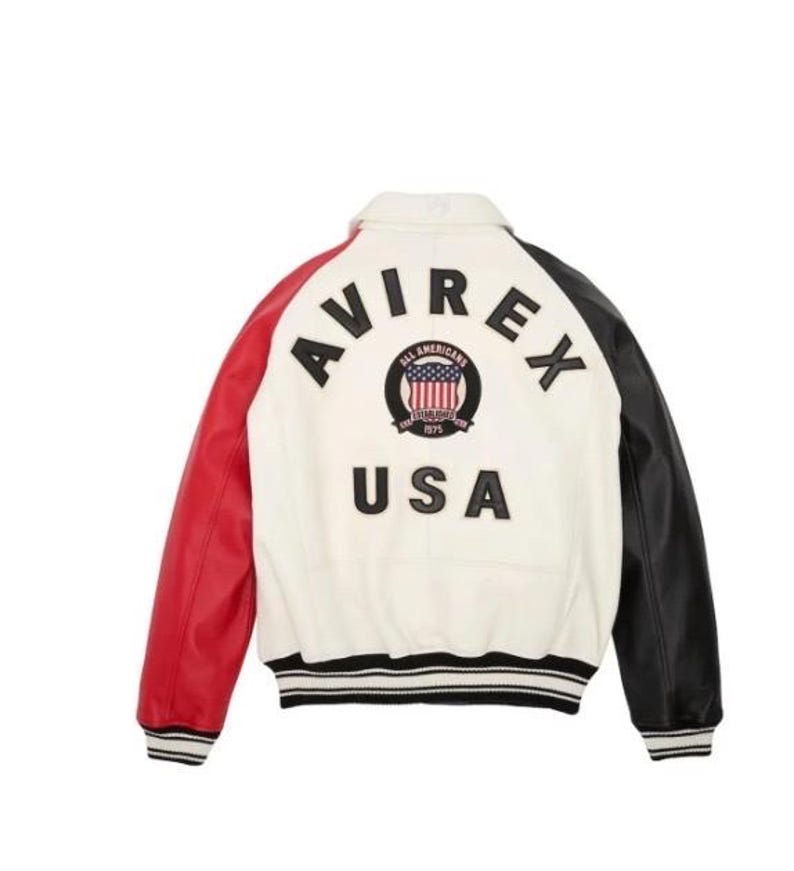 Op de afbeelding: Een varsity-jack met een wit achterpaneel, een rode rechtermouw en een zwarte linkermouw. De achterkant toont de zwarte tekst AVIREX, een ronde patch met een Amerikaans vlag-embleem en de tekst ALL AMERICANS ESTABLISHED 1975, en de tekst USA.