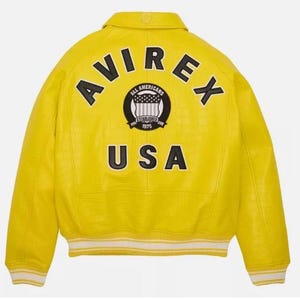 K&ouml;nnte beinhalten: Eine leuchtend gelbe Collegejacke mit strukturierter Oberfl&auml;che, klassischem Kragen sowie gestreiften Rippb&uuml;ndchen an &Auml;rmeln und Saum. Auf der R&uuml;ckseite befinden sich der schwarze Schriftzug AVIREX, ein rundes Emblem und der Text USA.