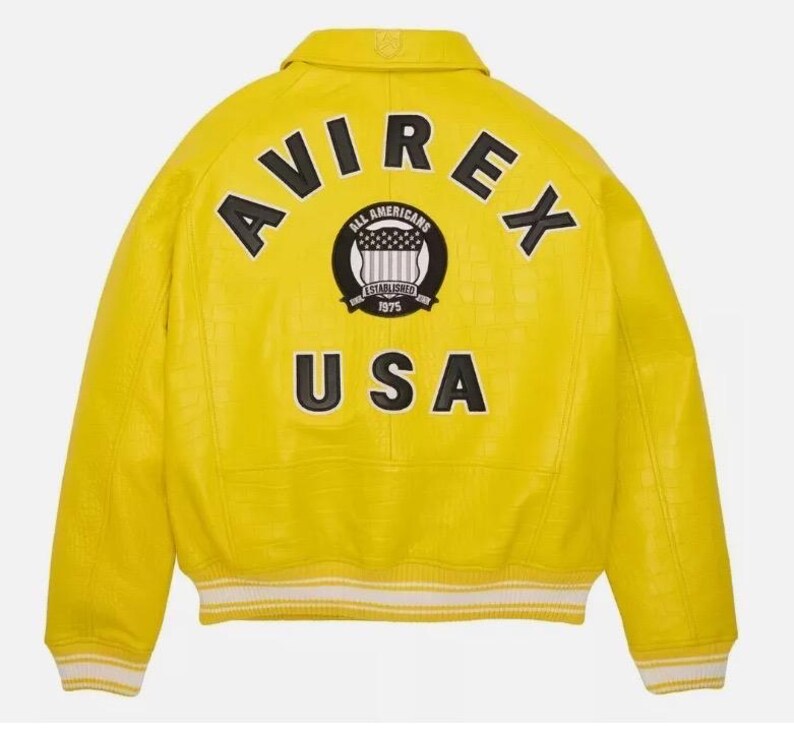 K&ouml;nnte beinhalten: Eine leuchtend gelbe Collegejacke mit strukturierter Oberfl&auml;che, klassischem Kragen sowie gestreiften Rippb&uuml;ndchen an &Auml;rmeln und Saum. Auf der R&uuml;ckseite befinden sich der schwarze Schriftzug AVIREX, ein rundes Emblem und der Text USA.