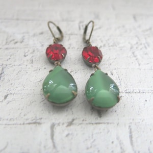 May include: A pair of vintage-style earrings. Each earring features a red, round gemstone at the top and a green, teardrop-shaped stone below. The metal has an antique bronze finish. The earrings are designed to dangle.