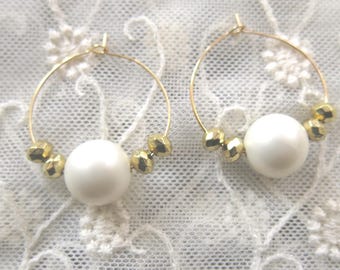 May include: A pair of gold-tone hoop earrings. Each earring features a large white pearl bead in the center, flanked by smaller, gold-colored geometric beads. The earrings are displayed against a white, lace-like background.