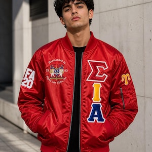 May include: A red satin bomber jacket with embroidered patches. The jacket features the letters "ΣIA" in white, yellow, and blue, along with the text "FA 20" and a crest. It has a zipper closure and a ribbed collar and cuffs.