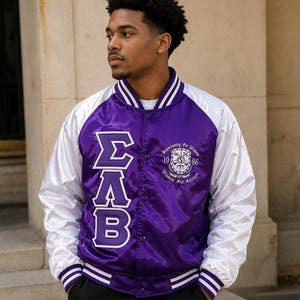 May include: A satin varsity jacket in purple and white. The jacket features the Greek letters ΣAB in white on the front, along with text and a crest. The sleeves are white, and the body is purple. The jacket has a button-up front and striped cuffs.