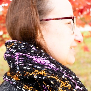 May include: A woman wearing a black and white patterned scarf with pink and orange accents. The scarf is knitted and has a loose, flowing style. The woman is wearing glasses and has brown hair.