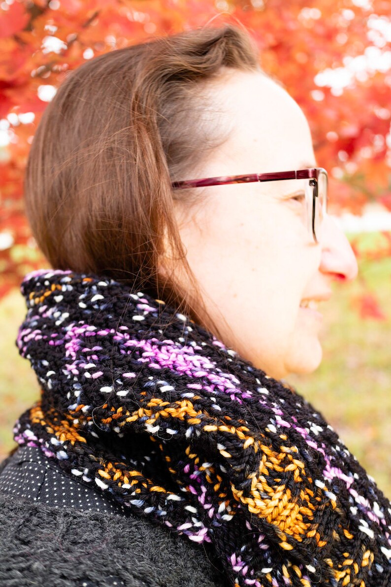 May include: A woman wearing a black and white patterned scarf with pink and orange accents. The scarf is knitted and has a loose, flowing style. The woman is wearing glasses and has brown hair.
