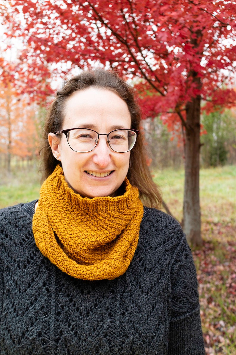 May include: A woman wearing a black sweater with a brown knitted infinity scarf. She is standing in front of a tree with red leaves.