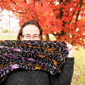 May include: A person wearing glasses and a black jumper holds a colourful knitted scarf in front of their face. The scarf has a black background with purple, orange, and white flecks. The person is standing in front of a tree with red leaves.