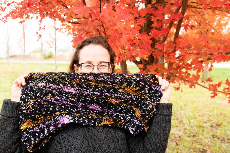 May include: A person wearing glasses and a black jumper holds a colourful knitted scarf in front of their face. The scarf has a black background with purple, orange, and white flecks. The person is standing in front of a tree with red leaves.