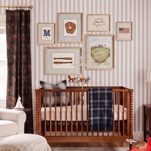 May include: A wooden crib with a dark blue plaid blanket and a gray plaid pillow, positioned against a wall with six framed baseball-themed prints, including a glove, bats, a stadium, and the text Long Live Boyhood and an American flag.