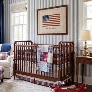May include: A dark wood crib with a red, white, and blue plaid quilt and matching bumper. A framed print of the American flag with the text God Bless America hangs above. A red toy airplane sits on the floor in front of the crib.