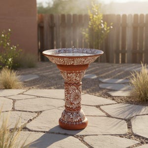 Puede incluir: Un bebedero para pájaros de terracota con base de pedestal. El cuenco y el pedestal están decorados con motivos florales blancos. El bebedero está al aire libre, con agua en el cuenco y un patio de piedra en primer plano.