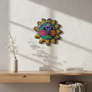 May include: A circular ceramic sun wall plaque featuring a central face with large eyes, a blue nose, and a green mouth. The sun is surrounded by yellow and blue rays, with pink and orange floral accents on the cheeks and forehead.