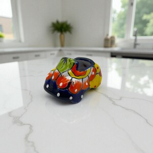 Puede incluir: Una pequeña maceta de cerámica pintada a mano con forma de zapato. La maceta presenta un diseño vibrante con colores azul, amarillo, rojo y verde. Lunares blancos acentúan la base azul. La maceta con forma de zapato es un artículo decorativo.