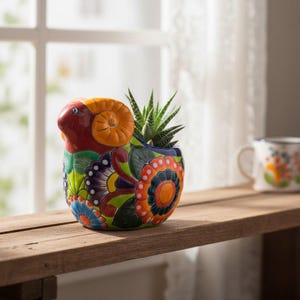 May include: A ceramic planter shaped like a ram with curled yellow horns, painted in vibrant red, green, blue, and orange floral patterns. A small green succulent plant with pointed leaves is potted inside the hollow back of the ram figure.