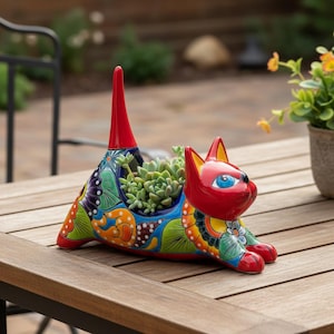 Op de afbeelding: Een keramische plantenbak in de vorm van een kat met een levendig, veelkleurig bloemen- en geometrisch patroon. De kat heeft een felrode kop, oren en een hoge, rechtopstaande staart, met grote blauwe ogen. Een kleine vetplant zit in het lichaam.