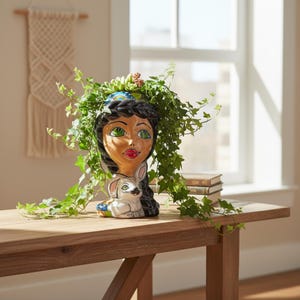 May include: A ceramic head-shaped planter featuring a painted face with green eyes, dark hair, and red lips, with a small white rabbit figure at the base. The planter is filled with a trailing green ivy plant and a small succulent on top.