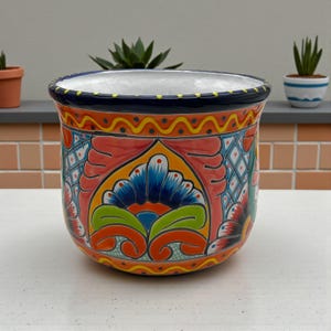 May include: A vibrant, hand-painted ceramic planter with a colorful floral design. The planter features a wide, cylindrical shape with a dark blue rim and a white interior. The exterior is adorned with intricate patterns in shades of red, orange, blue, and green.
