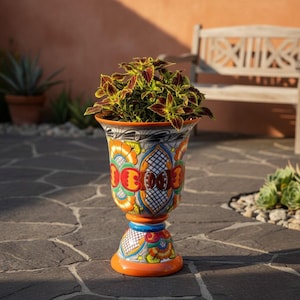 May include: A tall, pedestal-style ceramic planter featuring a vibrant, hand-painted Talavera-inspired pattern with orange, yellow, blue, and black floral and geometric motifs. A leafy coleus plant with variegated green and maroon foliage sits inside.