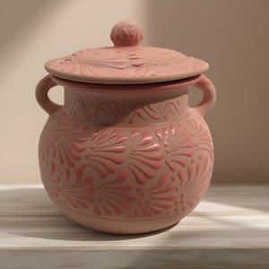 Puede incluir: Un jarrón de cerámica color terracota con tapa y dos asas. El jarrón y la tapa están decorados con un motivo floral en relieve en un tono rosa más oscuro. El jarrón está sobre una superficie de madera blanca.