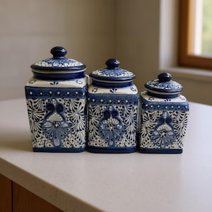 May include: Three ceramic kitchen canisters with lids. The canisters are square with rounded corners and feature a blue and white floral design. The lids are dark blue with a white floral pattern. The canisters are arranged in a row on a light-colored countertop.