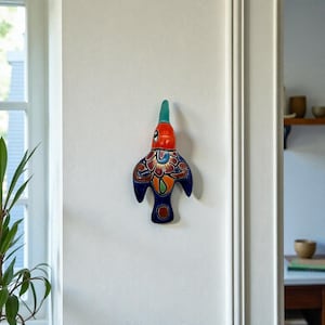 Puede incluir: Un colgante de pared de cerámica con forma de pájaro colorido. El pájaro tiene la cabeza roja, el cuerpo azul y el pico turquesa. El cuerpo está decorado con motivos florales en tonos naranja, amarillo y azul. Está montado en una pared blanca.