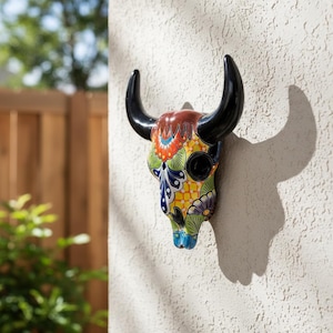 Puede incluir: Una decoración de pared con forma de cráneo de toro hecha de cerámica, con un patrón floral y geométrico pintado a mano en colores vivos. La pieza tiene cuernos negros brillantes y un acabado pulido, con detalles en naranja, amarillo, azul y verde.