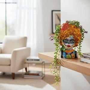 May include: A ceramic planter shaped like a human head with vibrant orange hair, intricate sugar skull face paint in blue, black, and red, and a colorful patterned collar, filled with trailing green succulent plants that drape over the sides.