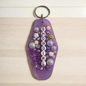 May include: A purple motel-style keychain with the word "SHOWGIRL" spelled out vertically in white letters. The keychain is decorated with small flowers, hearts, smiley faces, and clear bubbles, attached to a silver key ring.