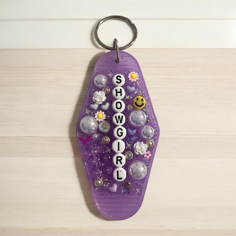 May include: A purple motel-style keychain with the word "SHOWGIRL" spelled out vertically in white letters. The keychain is decorated with small flowers, hearts, smiley faces, and clear bubbles, attached to a silver key ring.