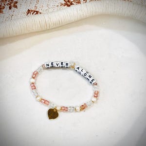 May include: A bracelet with the words "NEVER ALONE" spelled out in white letter beads. The bracelet features clear, pink, and gold beads, and a small gold heart charm.