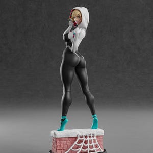 Może przedstawiać: Figurka żeńskiej postaci w czarno-białym body z kapturem. Postać stoi na ceglanej podstawie z białym wzorem pajęczyny. Turkusowe szpilki.