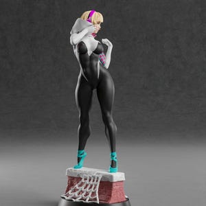 K&ouml;nnte beinhalten: Figur einer Superheldin in einem schwarz-wei&szlig;en Anzug, die auf einer Ziegelstruktur steht. Die Figur hat blondes Haar, ein rosa Stirnband und t&uuml;rkisfarbene High Heels. Der Sockel hat ein Spinnennetz-Design.
