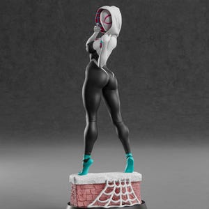 K&ouml;nnte beinhalten: Figur einer Superheldin in einem schwarz-wei&szlig;en Body, t&uuml;rkisfarbenen Abs&auml;tzen und einer wei&szlig;en Kapuzenmaske. Die Figur steht auf einem ziegelartigen Sockel mit einem wei&szlig;en Spinnennetz-Design. Der Hintergrund ist einfarbig grau.