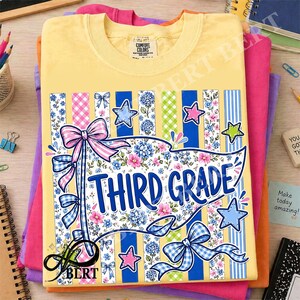 Könnte beinhalten: Ein hellgelbes T-Shirt mit einer Grafik eines Wimpels und dem Text THIRD GRADE in blauen Buchstaben. Das Design enthält vertikale Streifen, Blumenmuster, blaue und rosa Schleifen sowie sternförmige Symbole in Blau, Rosa und Grün.