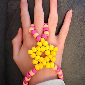 Op de afbeelding: Een handgemaakte kralenarmband en handstuk in de vorm van een ster. De armband heeft roze, oranje en gele kralen. De ster is gemaakt van gele kralen. De armband is verbonden met een ring die om de middelvinger gaat.