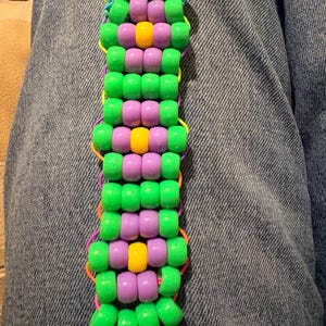 Könnte beinhalten: Ein handgefertigtes Kandi-Armband mit grünen, lila und gelben Perlen. Das Armband besteht aus einer Reihe farbiger Perlen, die mit elastischem Faden aufgereiht sind. Das Design umfasst abwechselnde Reihen aus grünen und lila Perlen, mit gelben Perlen in der Mitte.