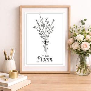 Puede incluir: Una impresión artística enmarcada con un dibujo lineal en blanco y negro de un ramo floral atado con un lazo. La palabra "Bloom" está escrita debajo del ramo. La impresión se muestra en un escritorio de madera con un jarrón de flores.