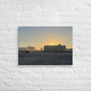 May include: A canvas print showing a beach scene at sunset. The image features a sandy beach with people and buildings silhouetted against the setting sun. The sky transitions from yellow to blue, creating a warm, inviting atmosphere.