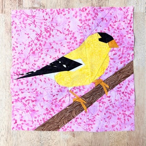 May include: A yellow and black paper pieced bird perched on a brown branch. The bird is surrounded by a pink and white floral pattern.