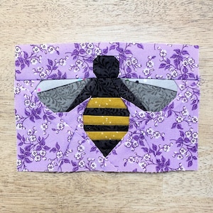 Könnte beinhalten: Ein lilafarbener Stoff-Bienenblock mit schwarzem Kopf, gelb-schwarzem gestreiftem Körper und weißem Blumenmuster im Hintergrund.