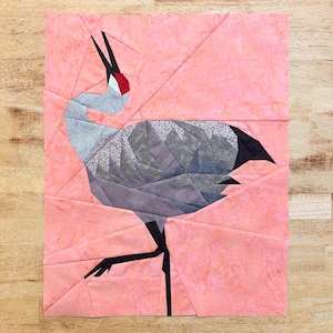 May include: A pink fabric background with a paper pieced crane design. The crane is made of various patterned fabrics in shades of gray, black, and red. The crane is standing on one leg with its head tilted back.