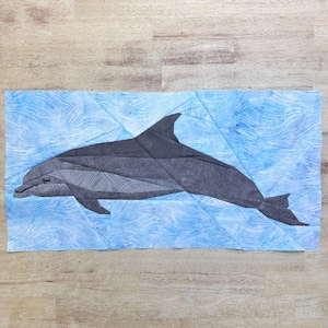 Puede incluir: Una obra de arte textil que representa un delfín gris en agua azul. El delfín está construido con formas geométricas, sobre un fondo azul con un patrón de olas. La pieza es probablemente un edredón o un tapiz de pared.