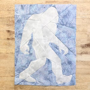 Può includere: Un ritaglio di carta bianco di Bigfoot che cammina su uno sfondo di tessuto con motivo blu e bianco.