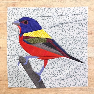 Peut inclure: Un bloc de courtepointe en patchwork coloré représentant un oiseau rouge, bleu, jaune et vert perché sur une branche. L'arrière-plan est un tissu bleu clair avec un motif de feuilles blanches.