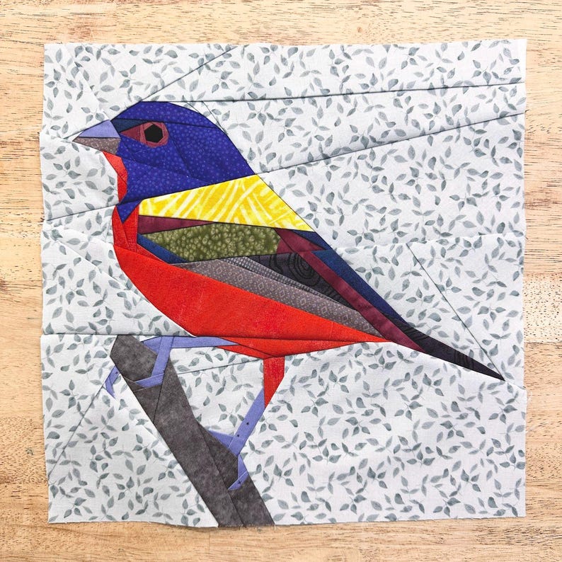 May include: A colorful patchwork quilt block featuring a red, blue, yellow, and green bird perched on a branch. The background is a light blue fabric with a white leaf pattern.