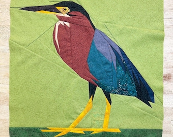 Peut inclure: Une œuvre d'art textile représentant un oiseau stylisé avec un long bec et des pattes jaunes. Le corps de l'oiseau est un mélange de tissus bordeaux, sarcelle et bleu, sur un fond vert. L'oiseau est posé sur une base verte.