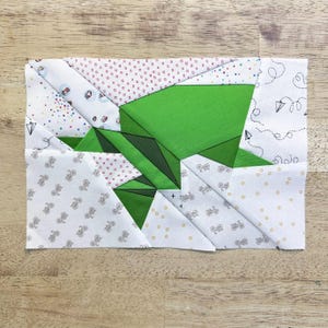 Pode incluir: Um bloco de colcha retangular com um design geométrico de um sapo de origami verde. O sapo está situado em um fundo de tecido branco com vários padrões, incluindo pequenas bicicletas, pontos e rabiscos extravagantes. O bloco está em uma superfície de madeira.