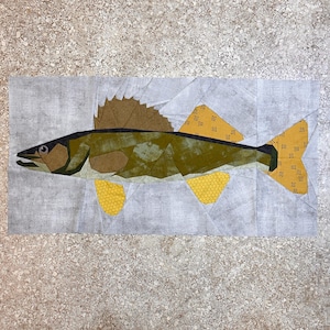May include: A patchwork quilt design of a walleye fish with brown, green, and yellow fabric pieces. The fish is swimming to the right.