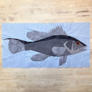 Puede incluir: Una obra de arte textil geométrica que representa un pez estilizado en tonos grises y negros, sobre un fondo azul claro. El pez tiene una aleta detallada y un ojo marrón. La obra de arte es probablemente un bloque de edredón o una creación textil similar.